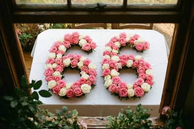 Roze en witte rozen vormen het getal 66 op een wit tafellaken, van bovenaf gezien binnen een houten raam- of deurkader met groen op de voorgrond en achtergrond.