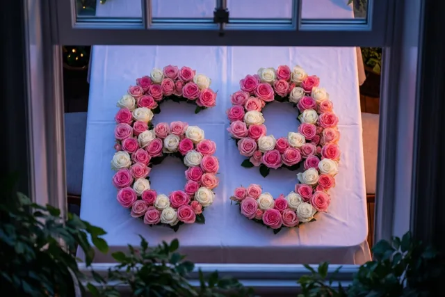 Van bovenaf door een raam is het getal 69 te zien, gevormd door roze en witte rozen op een wit tafelkleed in zacht, warm licht.
