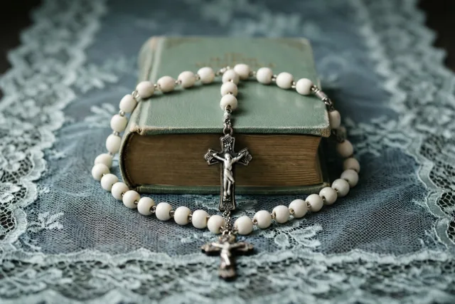 Een witte rozenkrans met een zilveren crucifix ligt op een klein gebedenboek met een groene kaft op een blauwe kanten doek.