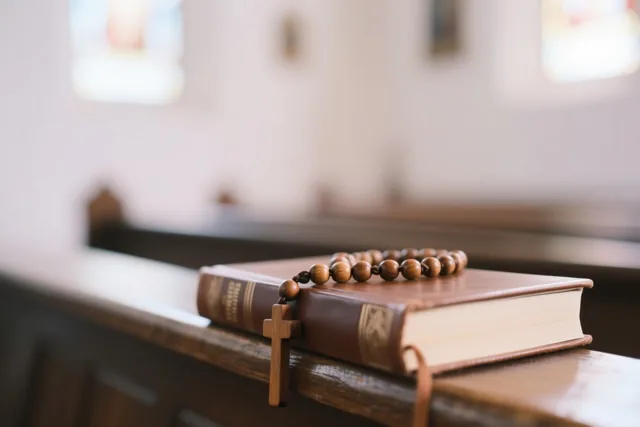 Een houten rozenkrans met kruis ligt op een gesloten leren gebedenboek op een houten kerkbank.