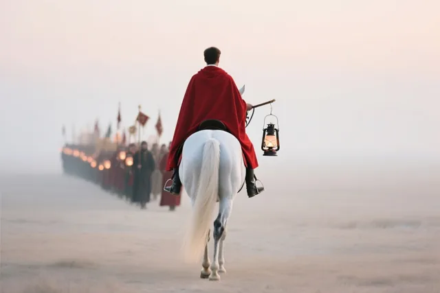 Een ruiter in een rode mantel op een wit paard leidt mensen met lantaarns door een mistig, open landschap in zacht en warm licht.