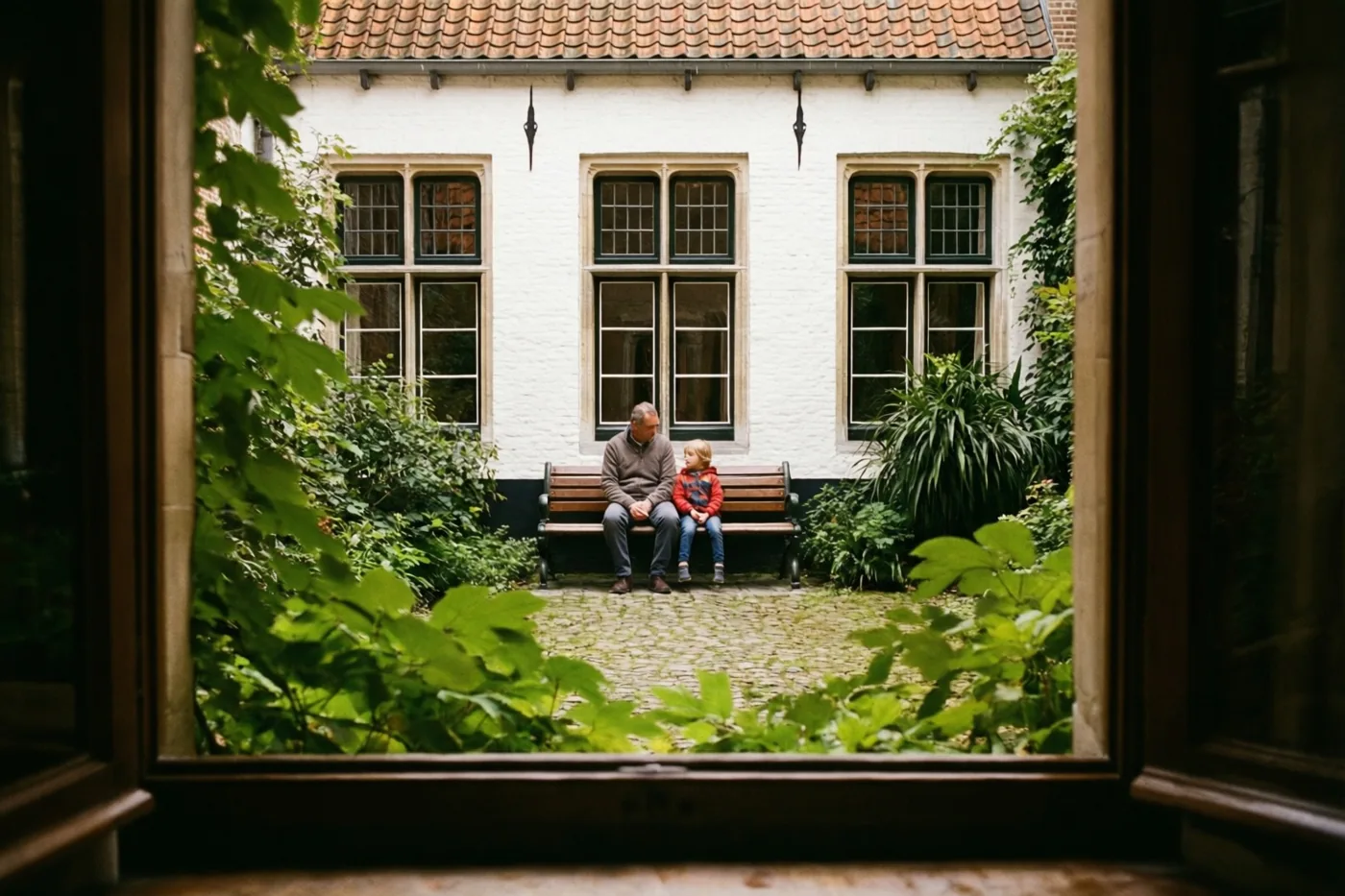 Een vader en kind zitten op een houten bank in een rustige binnenplaats, omringd door groen gebladerte en witte muren in zacht daglicht.