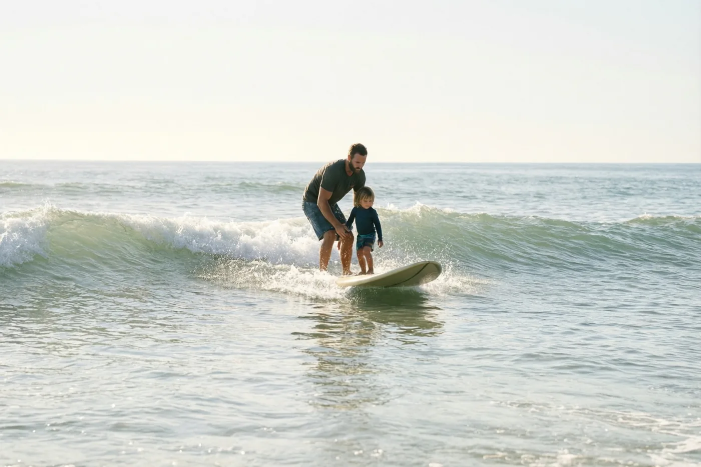 Een vader en zijn jonge kind surfen samen op een kleine golf in blauwgroen zeewater, van een afstand gezien onder een bleke hemel.