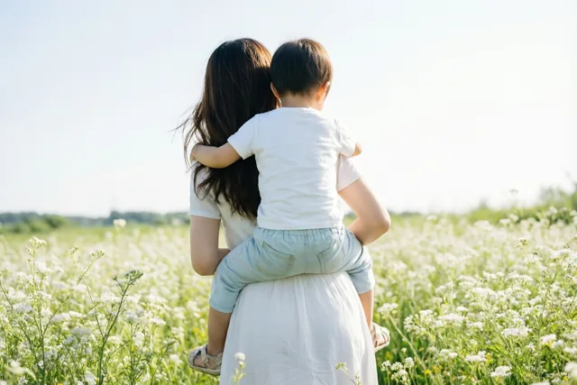 Een moeder met lang donker haar draagt een klein kind op haar rug door een veld witte wilde bloemen onder een heldere hemel, van achteren gezien.