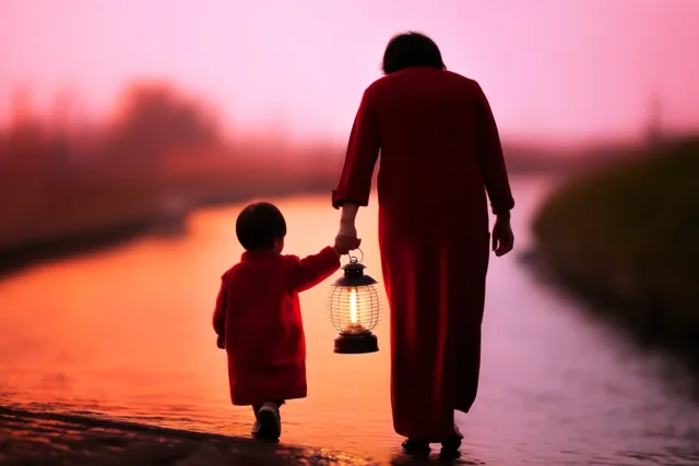 Een ouder en een klein kind lopen van de kijker weg hand in hand langs een kanaal met een brandende lantaarn, onder een roze en oranje zonsondergang.