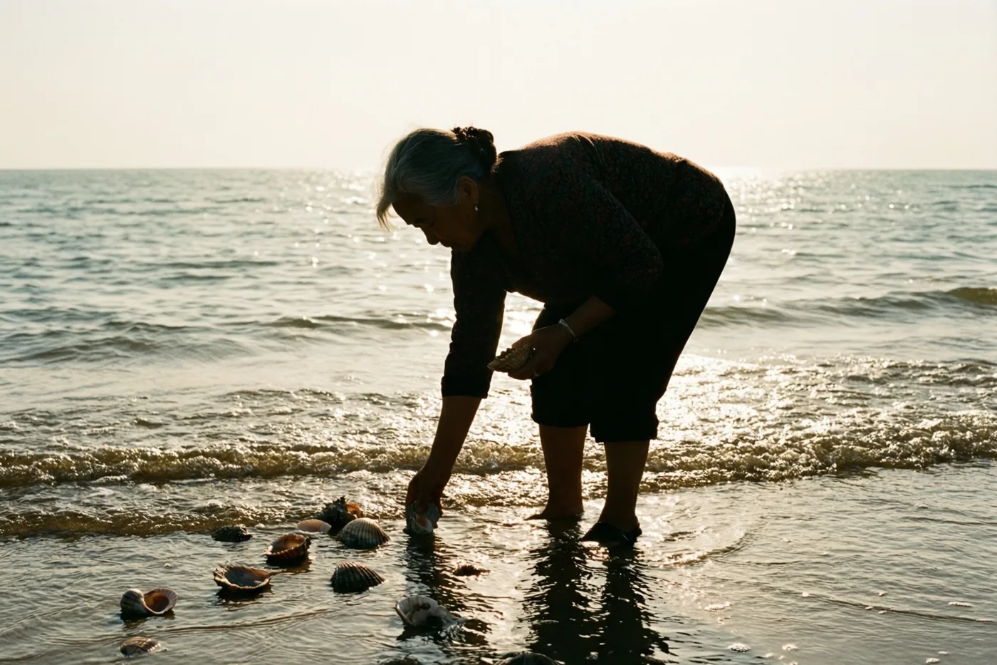 Het silhouet van een oudere vrouw buigt zich bij laagwater aan de waterlijn om schelpen te verzamelen, terwijl warm, goudkleurig licht op het water weerkaatst.