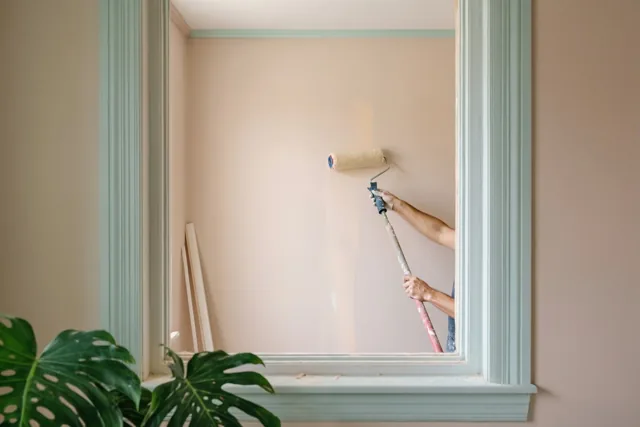 Door een lichtgroen raamkozijn is iemand met een verfroller te zien in een kamer met perzikkleurige muren en een groot monsterablad op de voorgrond.