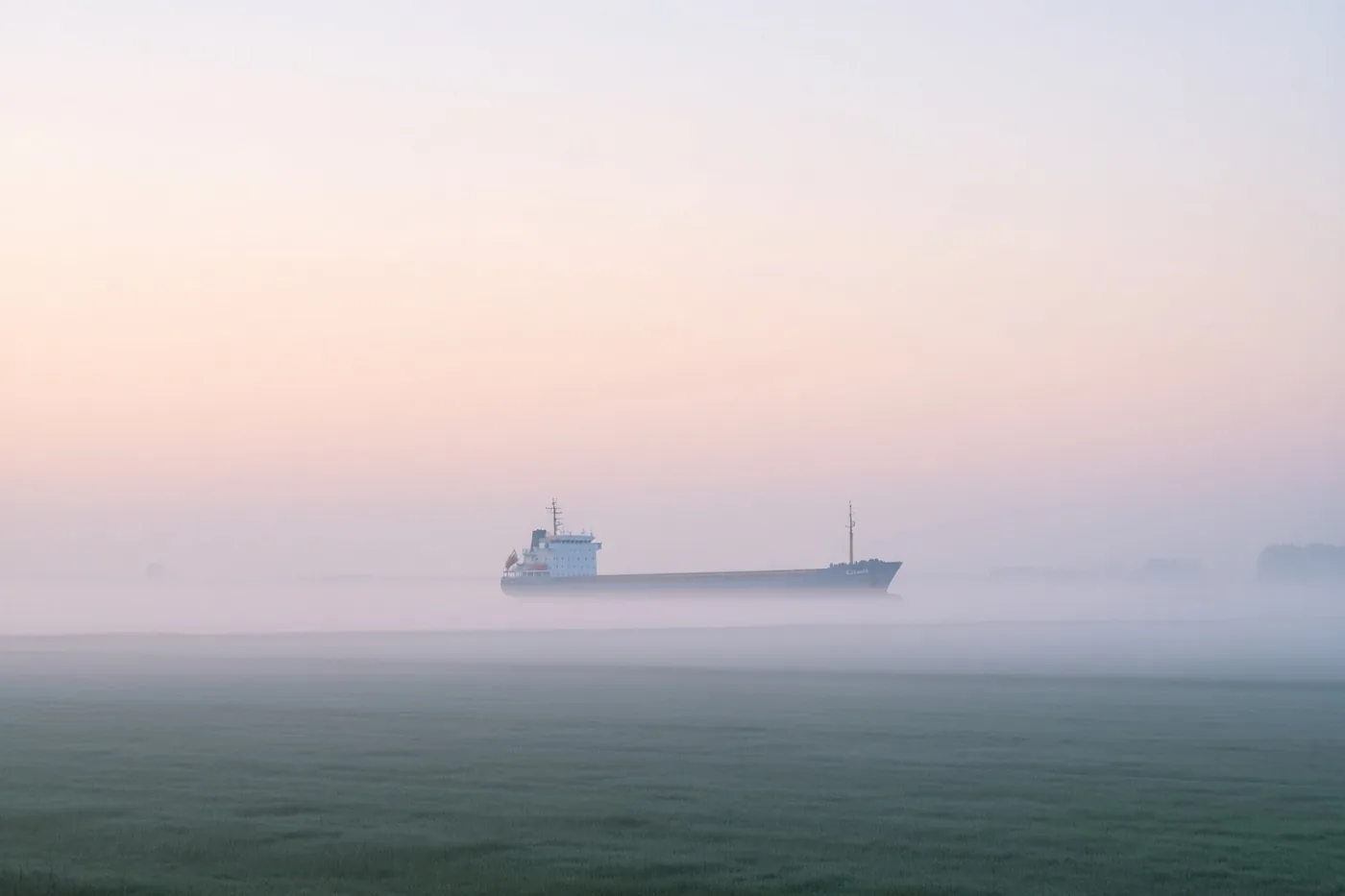 Een groot schip is in de verte deels verborgen door dichte mist boven rustig water bij zonsopgang, met een roze en lichtblauwe lucht en een groen veld vooraan.