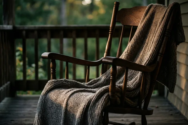 Een houten schommelstoel met een gebreid deken staat op een houten veranda, met een onscherpe groene tuin op de achtergrond in warm, zacht licht.