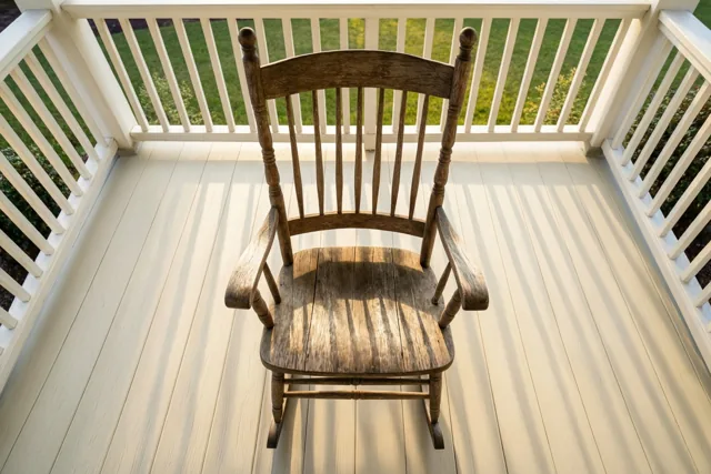 Een verweerde houten schommelstoel staat op een witte houten veranda met witte leuningen, met gras en struiken erachter in de namiddagzon.