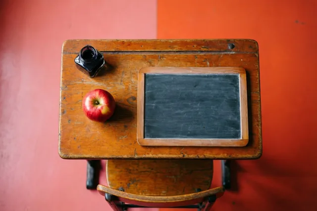 Van bovenaf zie je een oude houten schoolbank met een lei, een rode appel en een inktpot, tegen een roze-oranje muur.