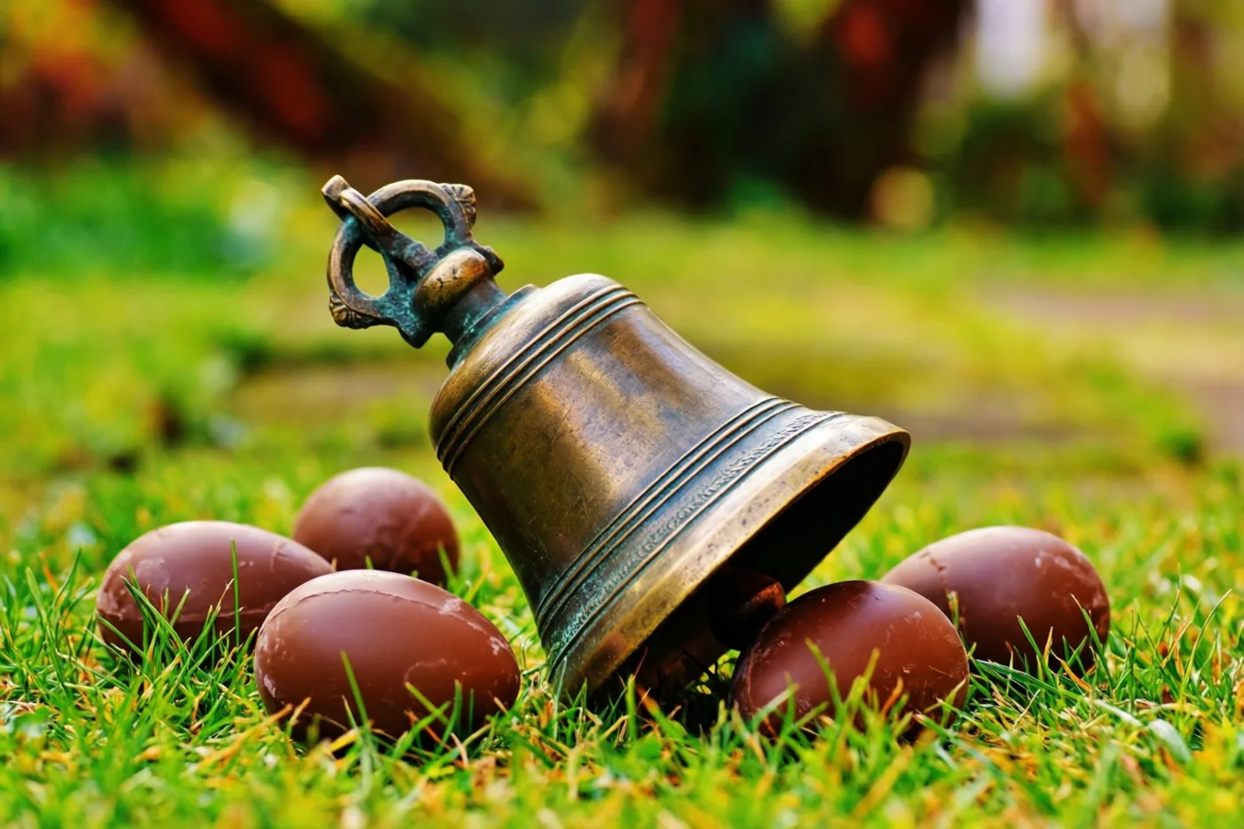 Een klein bronzen klokje ligt schuin in frisgroen gras tussen gladde chocolade-eieren bij natuurlijk licht.