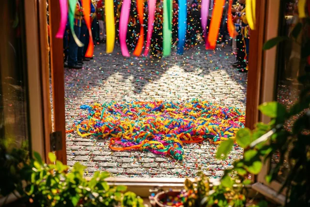 Door een houten deuropening zijn kasseien met kleurrijke serpentines en confetti te zien, met hangende serpentines erboven en groen op de voorgrond in fel zonlicht.