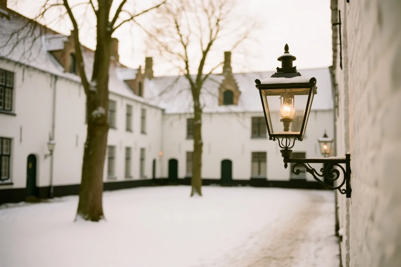 Een sierlijke brandende lantaarn staat op de voorgrond van een besneeuwd Brugs begijnhof met witte gebouwen en kale bomen in zacht, warm licht.