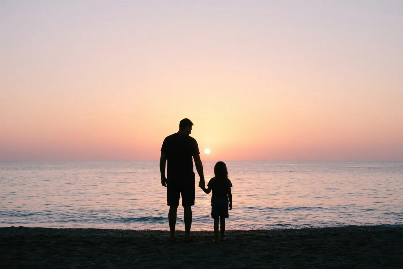 De silhouetten van een vader en kind staan hand in hand op het strand bij een zonsondergang boven zee, onder een oranje en roze lucht.