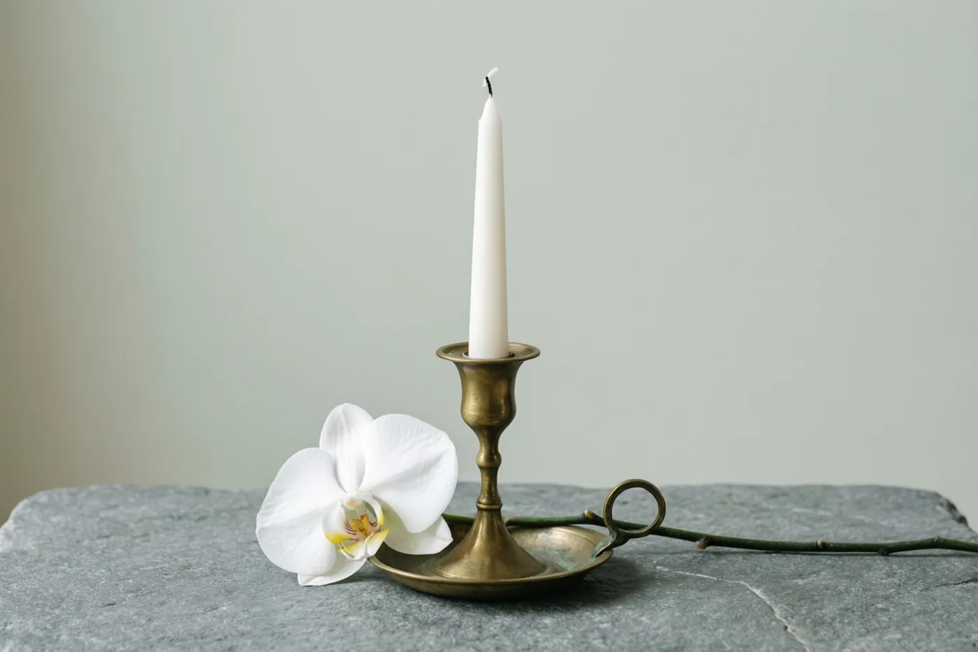 Een messing kandelaar met een slanke witte kaars staat naast een witte orchideebloem op grijze steen, in zacht licht en gedempte tinten.