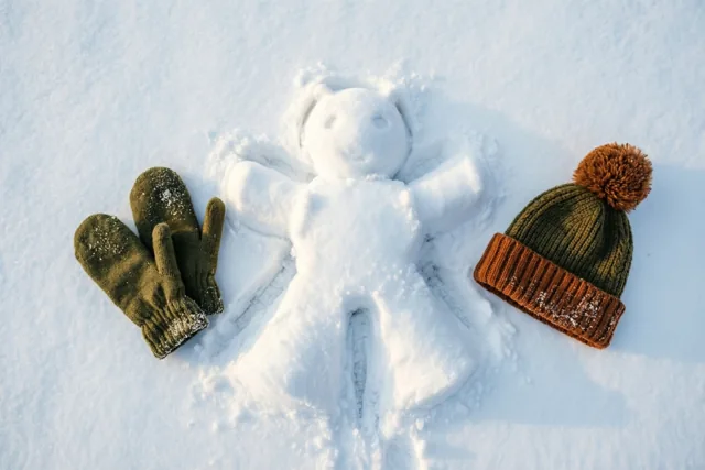 Een sneeuwengel ligt afgetekend in verse witte sneeuw, met groene wanten en een groenbruine pomponmuts ernaast in fel zonlicht.