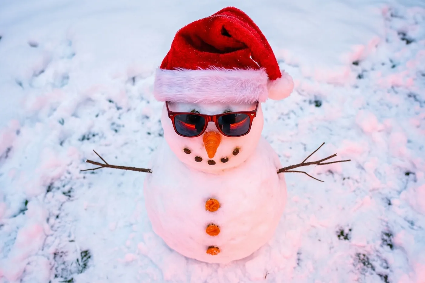 Een sneeuwman met een rode kerstmuts, rode zonnebril, wortelneus en oranje knopen staat op een besneeuwd gazon.