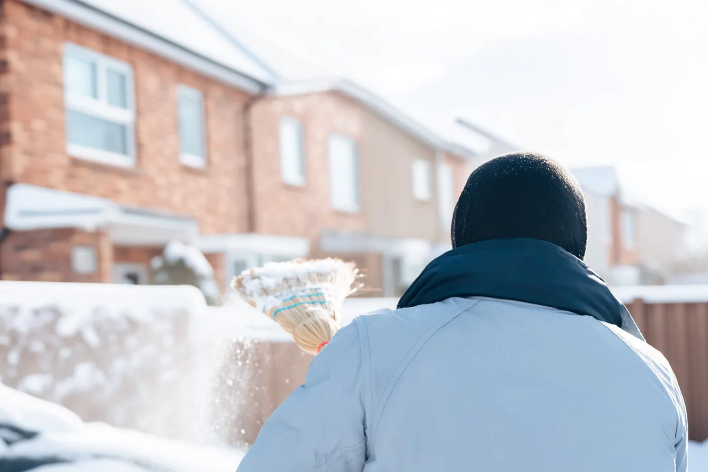 Een persoon met een lichte jas en donkere muts veegt, van achteren gezien, sneeuw met een bezem op een besneeuwd erf bij een bakstenen rijhuis onder een lichte, bewolkte hemel.