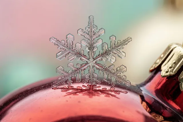 Een fijne sneeuwvlok rust op een glanzende rode glazen kerstbal, met een wazige achtergrond in pastelroze en blauwgroen.