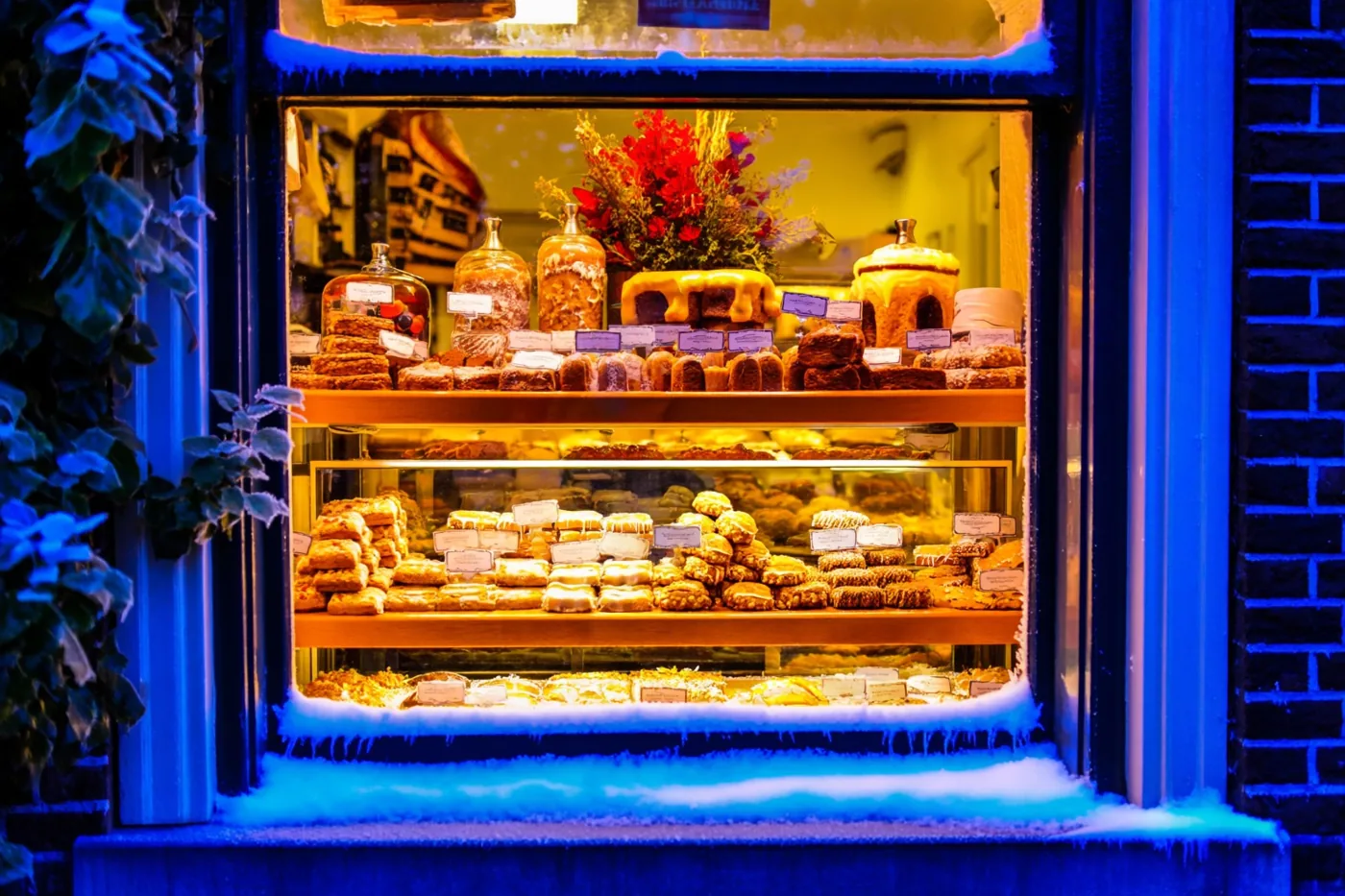 Een warm verlichte bakkersetalage toont rekken speculaas en marsepeingebak, met rijp en sneeuw op het raamkozijn en de vensterbank in blauw avondlicht.