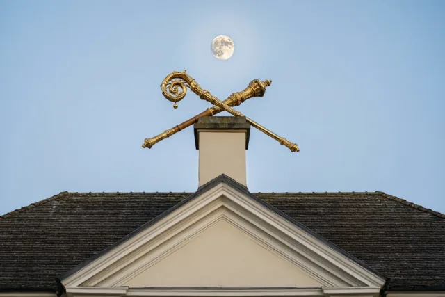 Een gouden bisschopsstaf staat boven op een schoorsteen, met roomkleurige bouwdetails en donkerbruine dakpannen onder een blauwe hemel met volle maan.