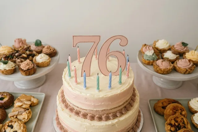 Een taart met verdiepingen, een grote papieren 76 en kleurrijke kaarsjes staat centraal op een tafel tussen schalen met gebak in zachte pasteltinten.