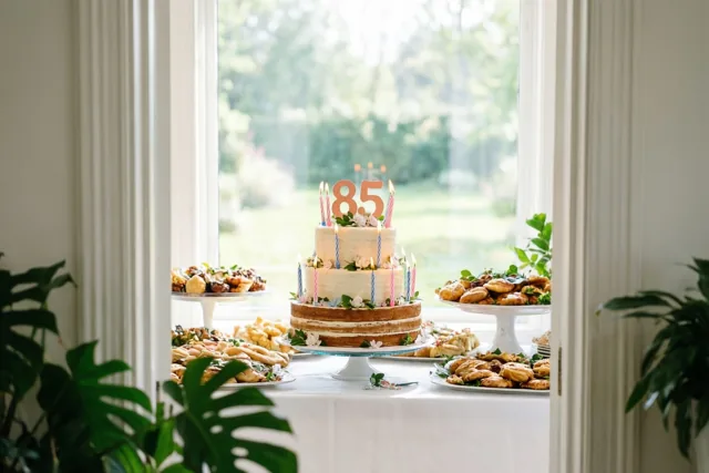 Een taart met verdiepingen, pastelkleurige kaarsjes en een papieren versiering met 85 staat op een buffet bij een raam, met een wit tafelkleed en groen.