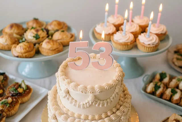 Een taart met meerdere verdiepingen, een papieren versiering met 53 en brandende kaarsjes staat op een buffet met pastelkleurige taartstandaards en allerlei gebak.