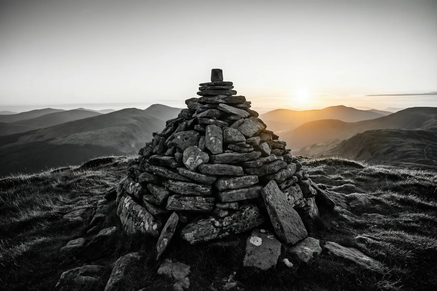 Een stapel stenen staat op een bergtop boven heuvels in zwart-wit met een warme gloed van de zonsopgang aan de horizon.