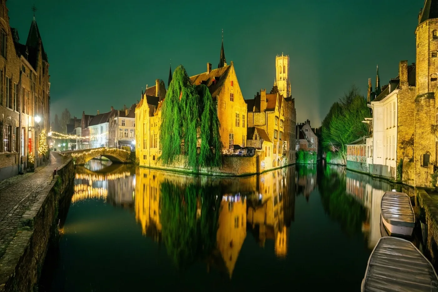 Oude gebouwen, groen getinte bomen en kerstlichtjes aan een stenen brug verlichten een nachtelijk tafereel langs het water in een Vlaams stadje.
