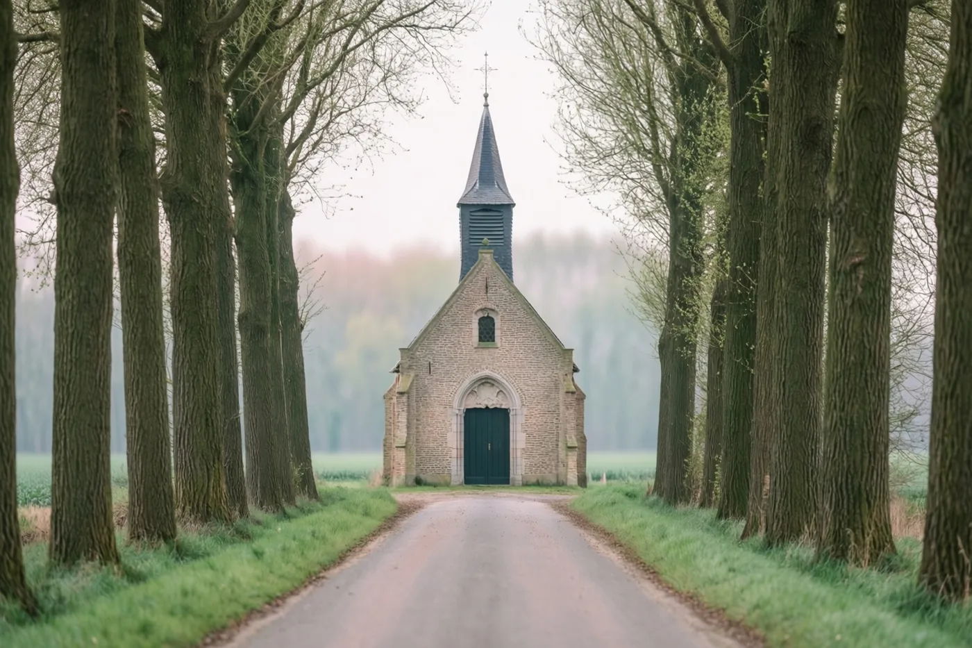 Een kleine stenen kapel met een donkere spits staat aan het einde van een landelijke laan tussen hoge kale bomen, in gedempte groene en grijze tinten.