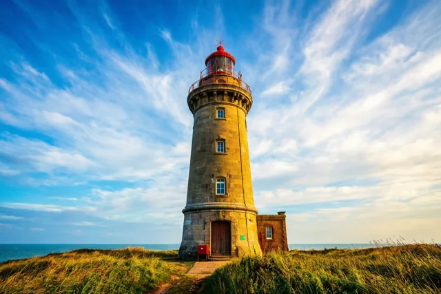 Een stenen vuurtoren staat op een met gras begroeide kustklif boven zee onder een blauwe hemel met sluierwolken in goudkleurig licht.