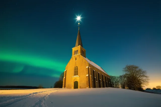 Een besneeuwde Vlaamse kerk staat onder een donkerblauwe sterrenhemel met groen poollicht en een heldere ster boven de toren, verlicht door warm kunstlicht.