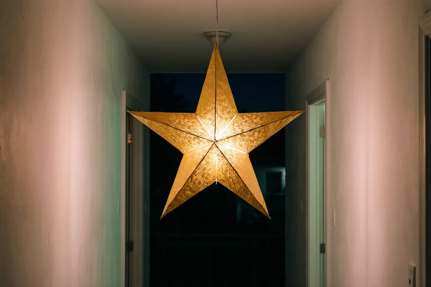 Een lichtgevende gouden papieren sterlantaarn hangt 's nachts aan het plafond van een smalle witte gang en verlicht de muren met warm licht.