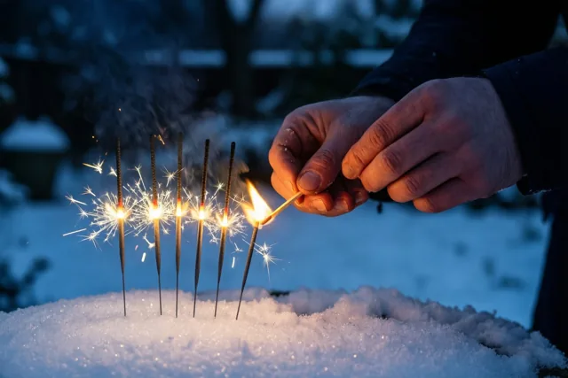 Handen strijken een lucifer aan bij rechtopstaande sterretjes in de sneeuw, met oranje vonken en vlammen tegen een donkerblauwe wintertuin.