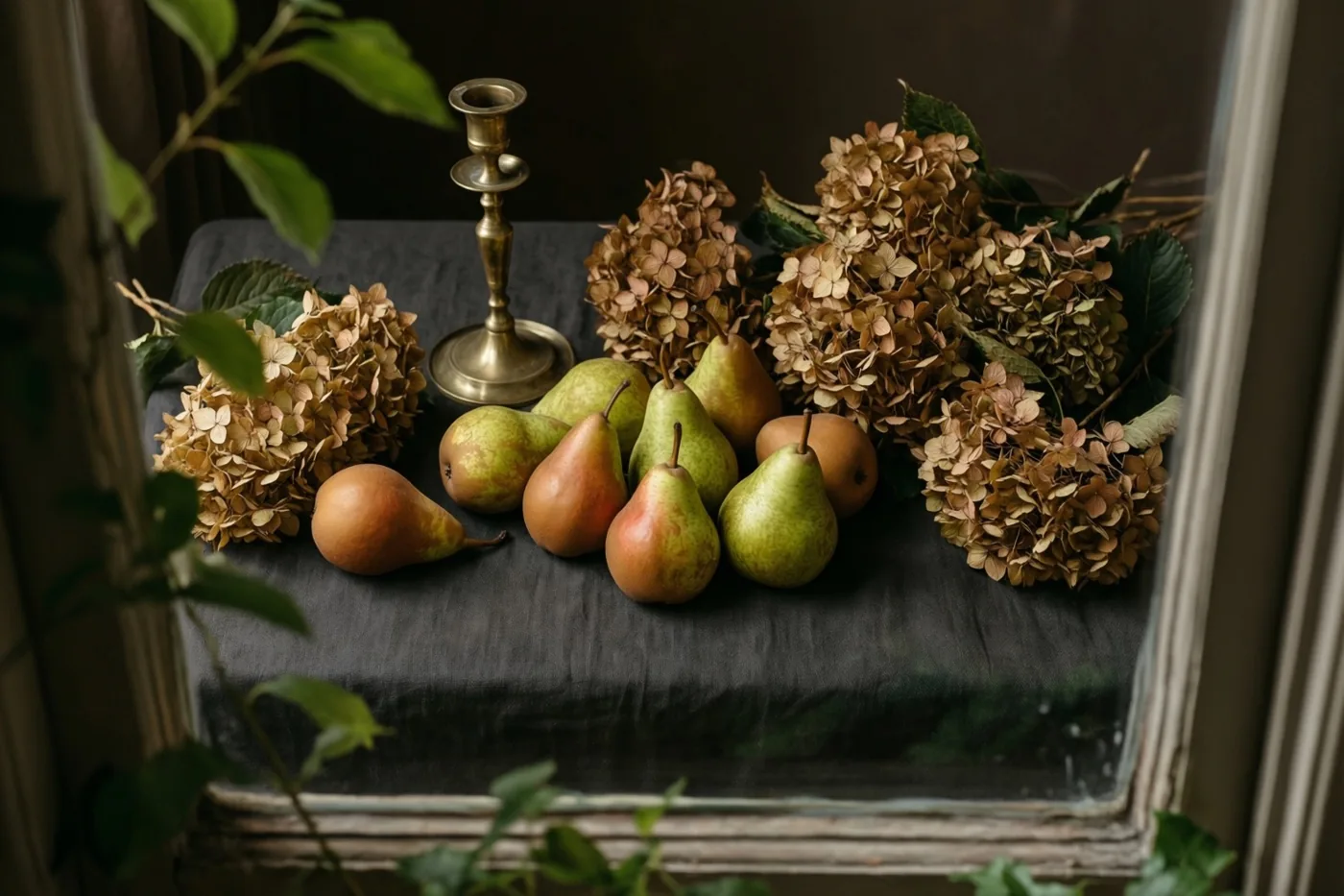 Peren, gedroogde hortensia's en een messing kandelaar staan op donker linnen in een omlijste spiegel, in zacht licht en aardetinten.