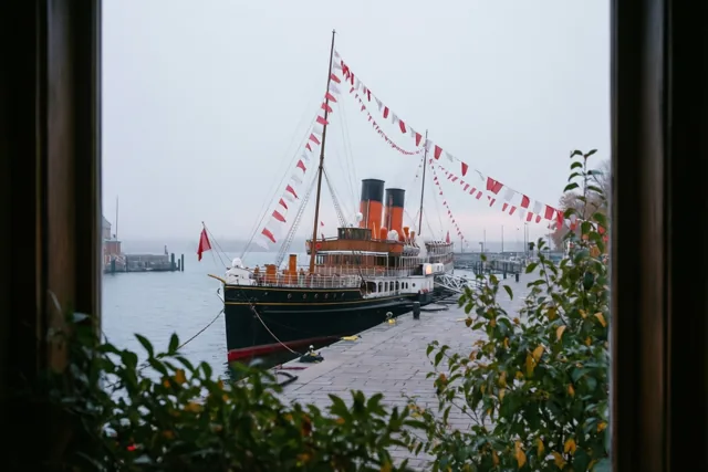 Door een raam met groen op de voorgrond is een aangemeerde stoomboot met oranje schoorstenen en rode en witte vlaggen te zien aan een kade op een grijze ochtend.