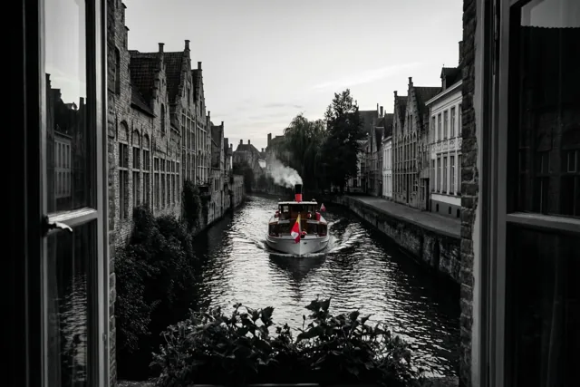 Door een raam is een stoomboot op een kanaal tussen oude Vlaamse gebouwen te zien, in zwart-wit met alleen kleur in de rode en witte vlaggen.