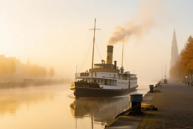 Een oude stoomboot met een gele schoorsteen vaart door een mistig kanaal naar een kade met herfstbomen en historische gebouwen in goudkleurig ochtendlicht.