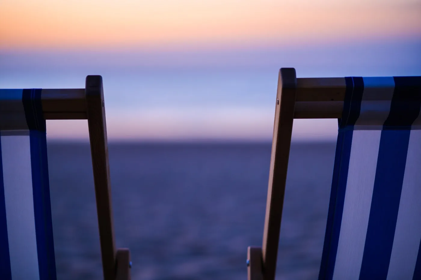 Twee blauw-wit gestreepte strandstoelen staan bij valavond naar zee gericht in zacht paars en oranje licht.