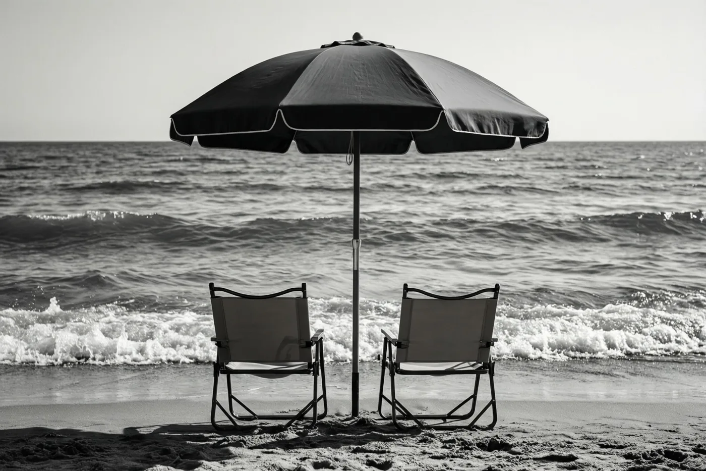 Op een zwart-witfoto staan twee klapstoelen onder een parasol aan de waterlijn, gericht naar de zee met zacht aanrollende golven.