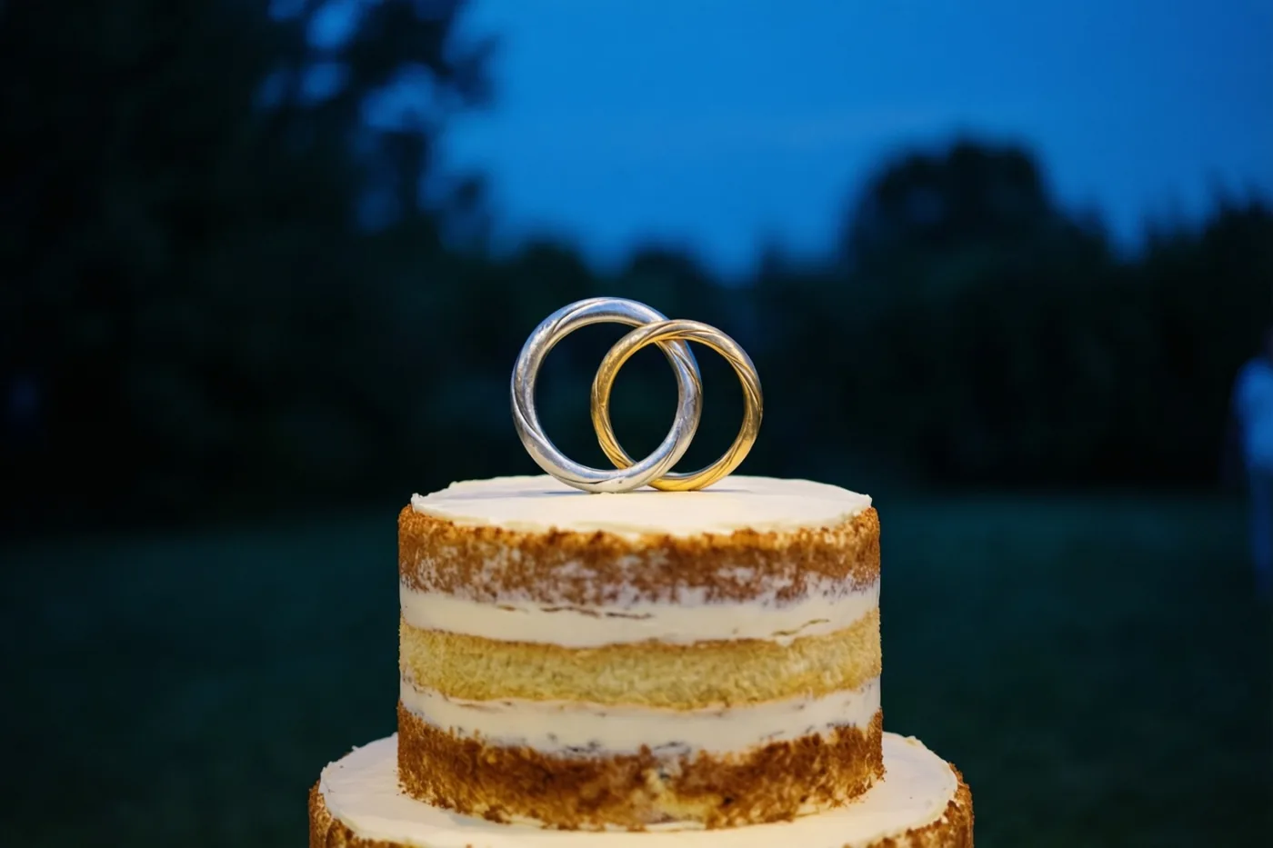 Een taart met twee verdiepingen, wit glazuur en zichtbare cakelagen staat buiten in de schemering, met bovenop een zilveren en een gouden ring die in elkaar haken, tegen een diepblauwe lucht en onscherp groen.