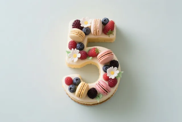 Een taart in de vorm van het cijfer 5 met bessen, macarons en eetbare bloemen staat tegen een effen lichtblauwe achtergrond.