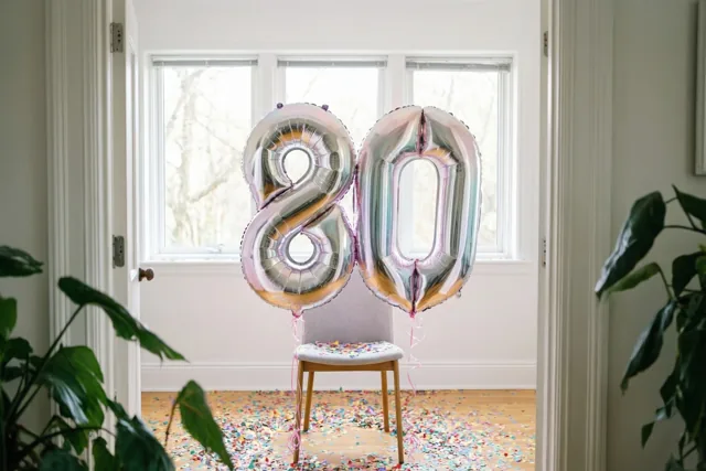 Zilveren en pastelkleurige cijferballonnen vormen 80 aan een stoel in een lichte kamer met confetti op de vloer, omlijst door deuropeningen en kamerplanten.