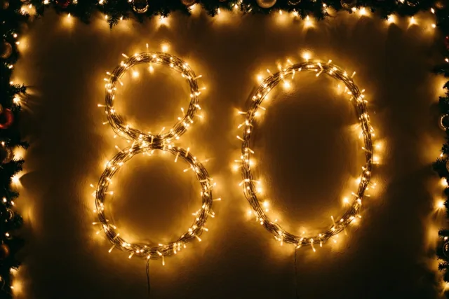 Warm goudgele lichtjes vormen het getal 80 tegen een donkere muur, omringd door decoratief groen en versieringen.