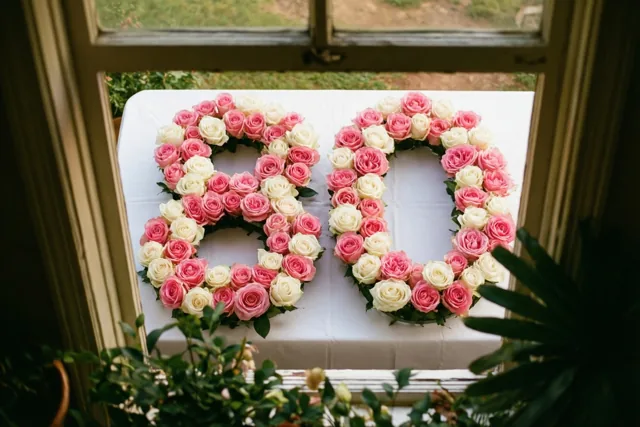 Door een raamkozijn zijn van bovenaf roze en witte rozen in de vorm van 80 op een wit tafelkleed te zien, met daglicht en groen buiten en vooraan.