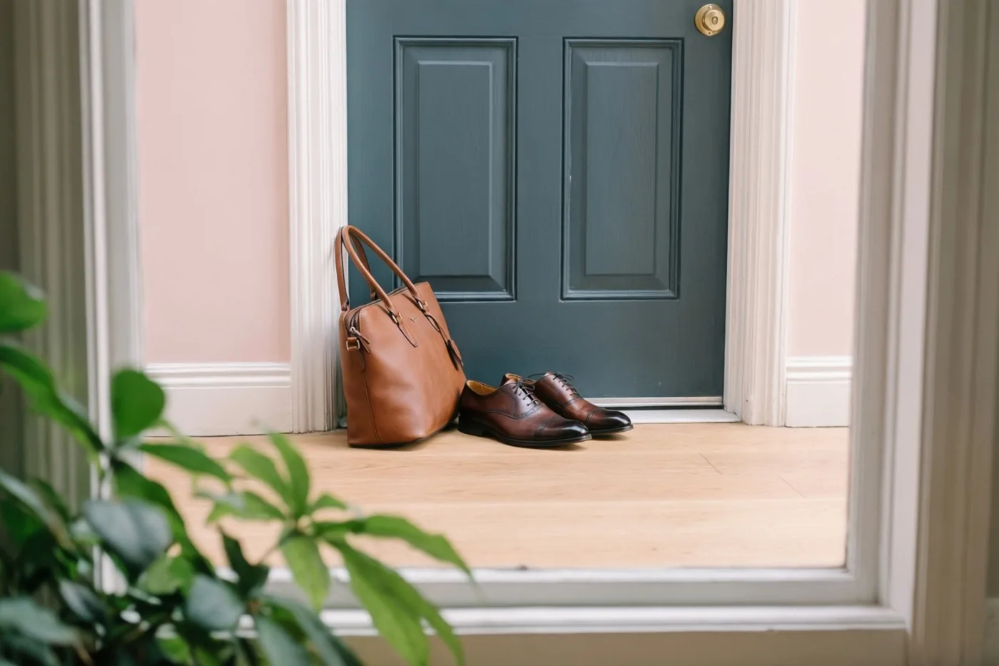 Een bruine leren tas en gepoetste bruine geklede schoenen staan op een lichte houten vloer voor een donkerblauwgroene deur tussen roze muren.