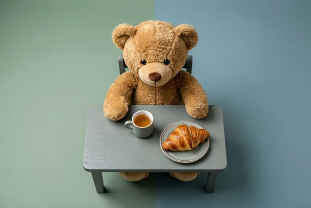 Een bruine teddybeer zit aan een klein grijs tafeltje met een kopje thee en een croissant op een bord, tegen een groen-blauwe achtergrond.
