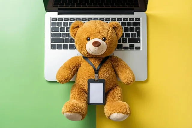Een bruine teddybeer met een onbedrukt halskoord zit op het toetsenbord van een laptop tegen een achtergrond met een groen en een geel vlak.